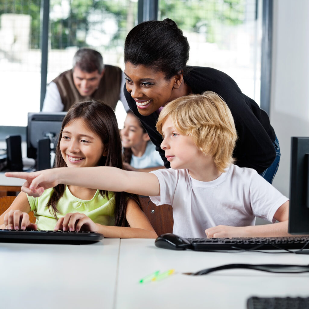 Teacher Assisting Boy Pointing On Computer In Lab